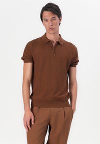 Polo castanho de manga curta com gola com fecho, punhos e bainha canelados. Usado com calças a condizer, exibindo uma palete de cores castanhas coesa.