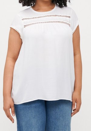 Woman wearing a white short-sleeve blouse with lace details across the chest and hemmed sleeves, paired with blue jeans.