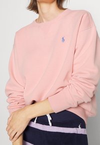 Sweatshirt rose clair avec des poignets et un col côtelés. Présente un petit logo brodé bleu. Tissu doux avec une coupe décontractée.