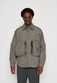 Lightweight, grey jacket featuring two large front pockets with black zippers, button closure, and a relaxed fit. Textured fabric with subtle sheen.