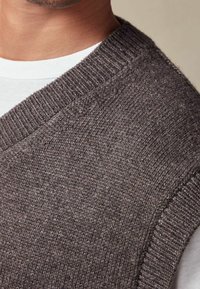 Gilet en tricot gris avec col et emmanchures côtelés, présentant un tissu texturé et doux. Porté par-dessus une chemise blanche en dessous.