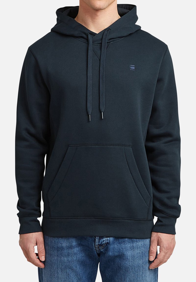 Sudadera de un oscuro color azul marino con capucha, cordones y un bolsillo frontal tipo canguro. Textura suave, ribete en el dobladillo y puños, logo en el pecho.