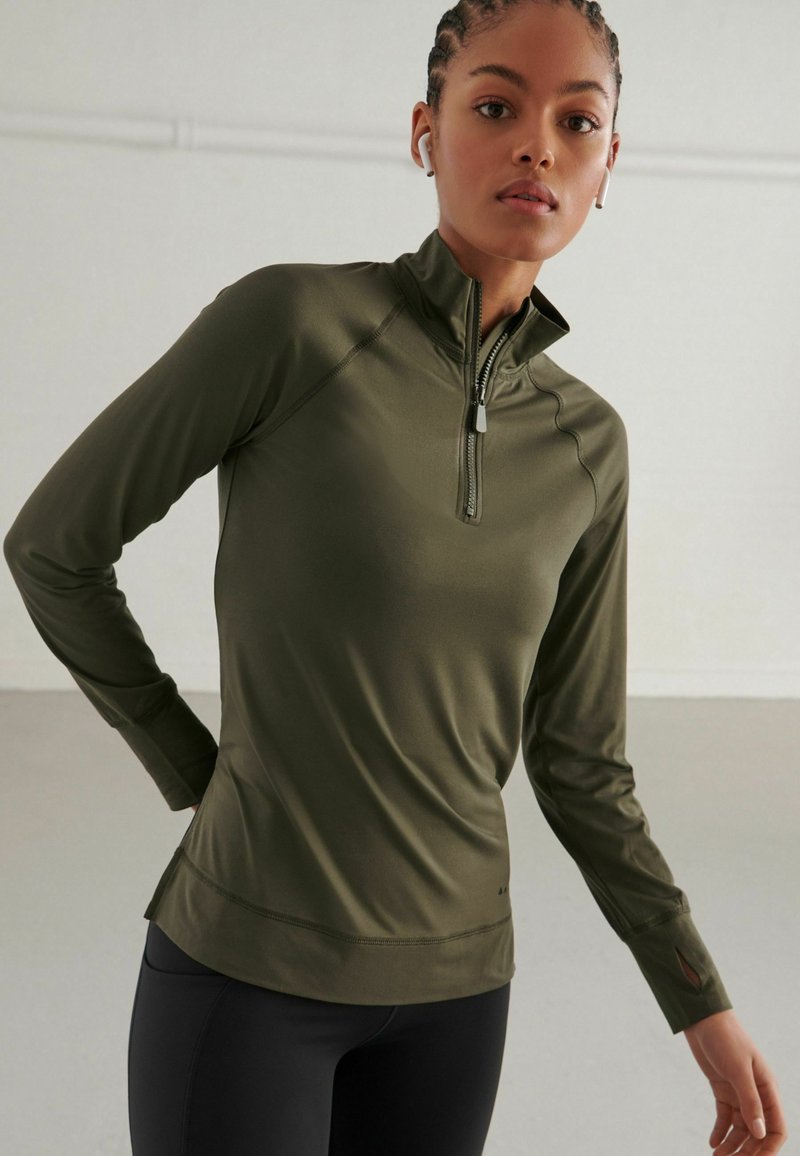 Tricou sport din material elastic, de culoare verde măslin, cu mâneci lungi și fermoar la jumătatea pieptului, cu croială ajustată, cusături plat și găuri pentru degete; fabricat dintr-un material neted și elastic.