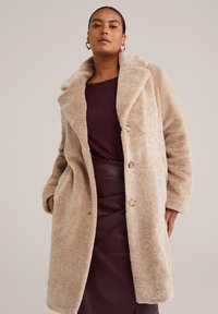Manteau en fausse fourrure beige avec grand col, fermeture à boutons et poches latérales. Porté sur un haut bordeaux et une jupe bordeaux foncé. Texture douce.