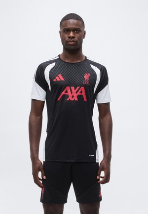 Homme portant un maillot d'entraînement noir et blanc du Liverpool FC avec des logos rouges et un short noir assorti, se tenant devant un fond uni.