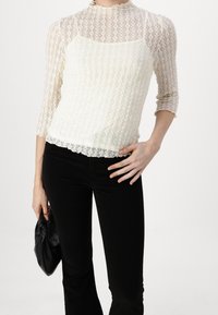 Top de encaje en crema con mangas tres cuartos, patrón texturizado, con borde en forma de ola, combinado con pantalones de campana negros y un clutch negro.