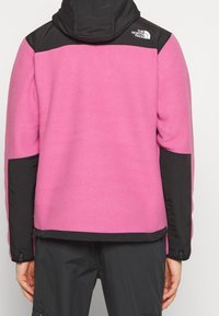 Chaqueta de forro polar en un brillante color rosa con detalles en nylon negro en los hombros y las mangas. Cuenta con capucha y un logo en la parte superior de la espalda.