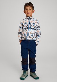 Chaqueta con estampado en blanco, azul y naranja; pantalones marinos con acentos en negro; botas grises y verdes; niño de pie.