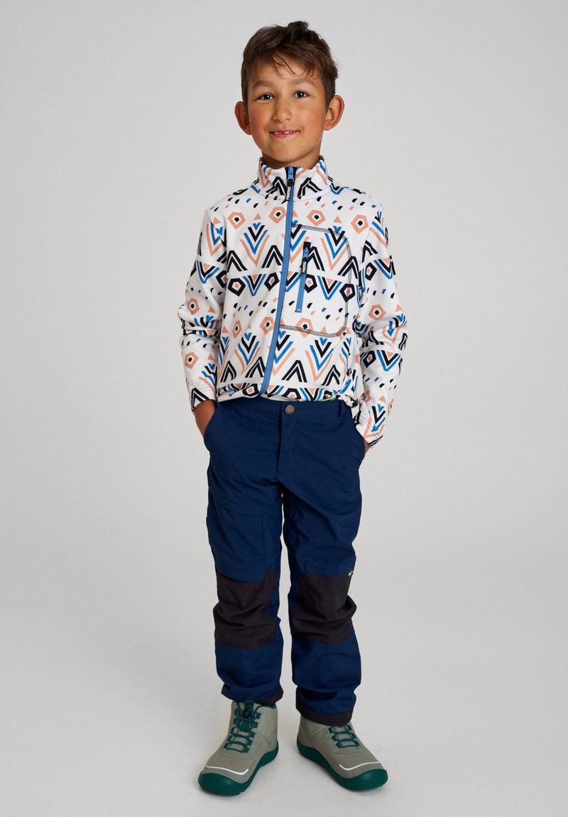 Chaqueta con estampado en blanco, azul y naranja; pantalones marinos con acentos en negro; botas grises y verdes; niño de pie.