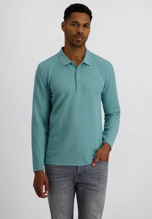 Mann steht da, trägt ein langärmeliges türkisfarbenes Polohemd und graue Jeans, eine Hand in der Tasche, vor einem schlichten Hintergrund.