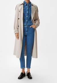 Beige trenchcoat över en jeansjacka och blå skinny jeans. Har knappdetaljer, en krage och svarta slip-on skor.