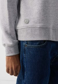 Felpa grigia con una finitura testurizzata, caratterizzata da un logo circolare sull'orlo, abbinata a jeans blu scuro. Primo piano del dettaglio della mano.