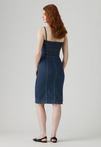 Vestido de mezclilla con una silueta ajustada, cuerpo fruncido y tirantes finos. Color: azul oscuro. Textura suave con detalles de costura visibles.