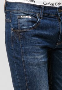 Jeans de mezclilla azul oscuro con un ajuste slim, que cuentan con un bolsillo frontal, un bolsillo trasero con branding y detalles de costura visibles.