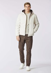 Giacca leggera beige con cappuccio, dotata di una fodera marrone, chiusura con zip e polsini elasticizzati. Abbinata a pantaloni marrone scuro e sneakers bianche.