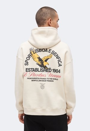 Personne portant un sweat à capuche crème avec un grand motif d'aigle et un texte sur le club sportif Benfica dans le dos.