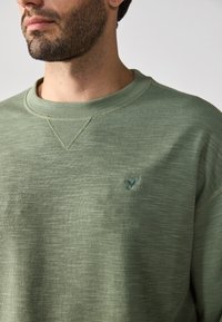 Svetlozelená mikina so štruktúrovaným, rebrovaným vzorom. Má okrúhly výstrih a malé vyšité logo na hrudi.