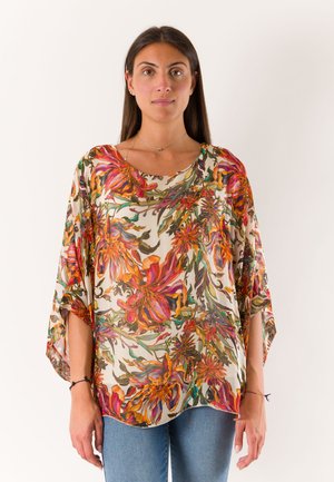 Donna con lunghi capelli scuri che indossa una camicetta larga con fantasia floreale nei colori arancione, rosa e verde, abbinata a jeans blu, in piedi su sfondo bianco.