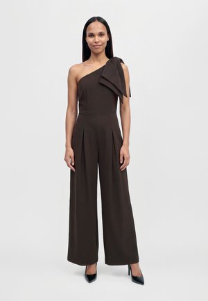 Une femme porte une combinaison marron foncé asymétrique à une épaule, ornée d’un grand nœud sur l’épaule et de jambes larges, associée à des chaussures noires à talons.