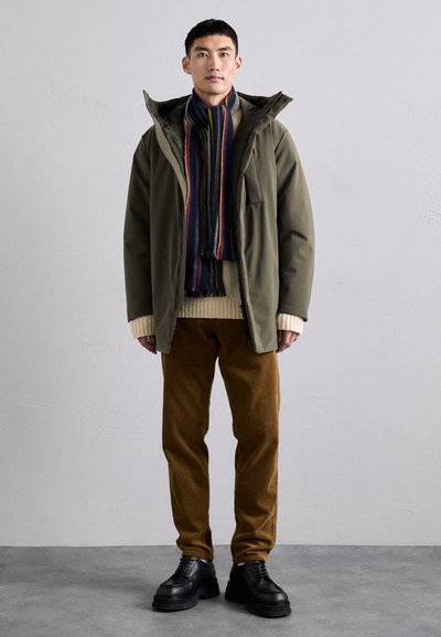 Giacca con cappuccio verde oliva, maglione in maglia crema, pantaloni in velluto a coste marroni, sciarpa a righe, scarpe nere chunky, in piedi contro uno sfondo neutro.