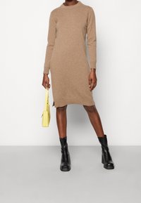 Robe beige en maille à manches longues, col rond et fentes latérales, associée à des bottines noires et un petit sac à main jaune.