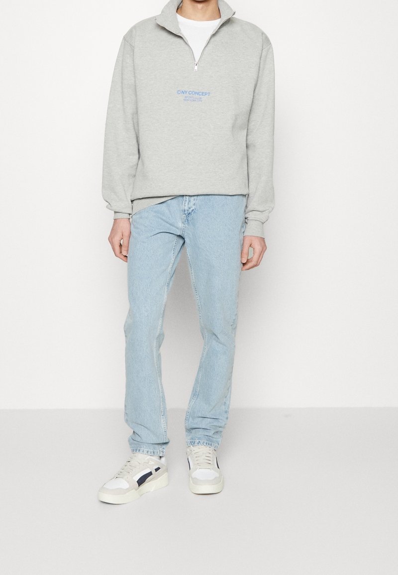 Personne portant un sweat-shirt gris clair à fermeture éclair quart de zip avec un logo bleu, un jean bleu clair, et des baskets blanches avec une bande foncée, debout devant un fond blanc.