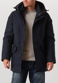 Marineblauwe donsjas met een ritssluiting aan de voorkant, grote capuchon, gestructureerde stof en zijzakken. Model draagt een lichte trui en jeans.