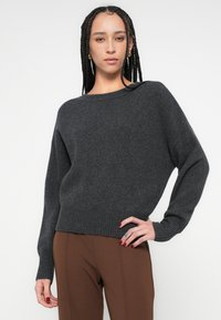 Tmavosivý pletený sveter so širokým výstrihom, opustenými ramenami a rebrovanými manžetami a lemom. Mäkká textúra, neformálny strih, v kombinácii s hnedými nohavicami.