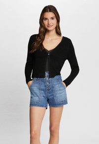 Cardigan nero a coste con chiusura a bottoni, abbinato a pantaloni costumi in denim blu a vita alta con ampie tasche e chiusura a cinque bottoni.