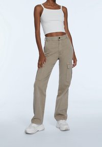 Beige cargobroek met zijzakken, gemaakt van katoen. Gepaarde met een witte cropped top en witte sneakers, staande tegen een lichte achtergrond.