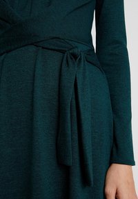 Tyrkysové pletené šaty s dlouhými rukávy, které mají zavinovací design a detail zauzleného pasu. Textura je měkká a texturovaná bez vzorů.