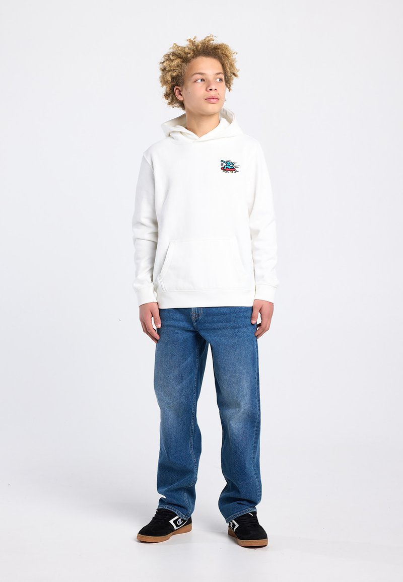 Tiener met krullend haar, gekleed in een witte hoodie met een kleine kleurrijke afbeelding, blauwe spijkerbroek en zwarte sneakers, staand tegen een effen witte achtergrond.