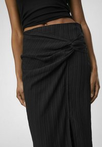 Jupe plissée noire avec un détail de nœud torsadé à la taille et une fente sur un côté. Le tissu texturé crée un motif subtil dans l'ensemble.
