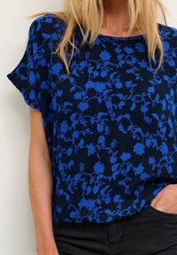 Mørkeblå top med korte ærmer, der har et blomstermønster i forskellige nuancer af blåt; ribbet tekstur og afslappet pasform.