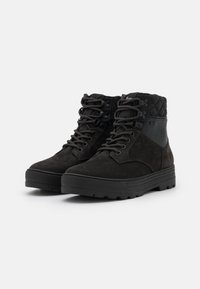 Botas de tobillo de ante negro con textura acolchada en el cuello, suela de goma y cordones redondos. Cuenta con un robusto hardware en forma de asa para un ajuste seguro.
