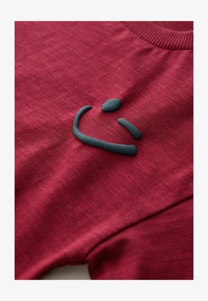 Piros textúrájú póló, elülső részén kiemelkedő sötétzöld hímzett mosolygós arc logóval, bordázott nyakkal.