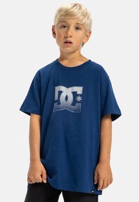 Modré bavlněné tričko s oversized střihem, které má na přední straně stříbrné texturované logo. Krátké rukávy a kulatý výstřih.