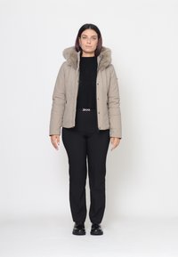 Giacca trapuntata beige con cappuccio foderato in pelliccia, chiusura con zip e due tasche laterali, abbinata a un maglione lavorato a maglia nero e pantaloni neri.