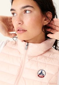 Pink quilted vest with high collar, zippered front, and round logo patch on chest. Smooth fabric texture and structured design.