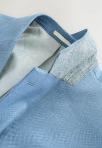 Detail světlomodrého texturovaného límce bundy se šedou filcovou vnitřní podšívkou a vzorovanou bílou látkou uvnitř.