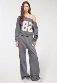 Młoda kobieta w luźnym szarym dresie z topem na jedno ramię i nadrukowanym numerem 82 na górze oraz na spodniach, stojąca na jednolitym tle.