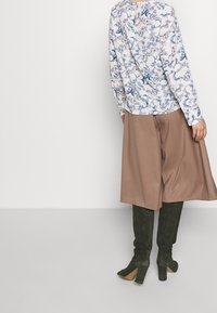 Donna che indossa una blusa floreale blu e bianca, una gonna larga marrone fino al ginocchio e stivali scamosciati verdi scuri al ginocchio, in piedi contro un muro bianco.