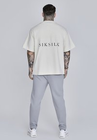 Prekračujúce krémové tričko s potlačou "SIKSILK" v čiernej na chrbte. V kombinácii so šedými joggers. Model ukazuje tetovanú ruku.