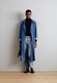 Trench coat in denim di tonalità azzurro, abbinato a un dolcevita nero e jeans a vita alta leggermente strappati. Calzature e borsa marroni.