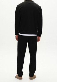 Mann, der eine schwarze Jacke und Hose trägt, darunter ein weißes Hemd, steht und ist von hinten auf einem schlichten weißen Hintergrund zu sehen.