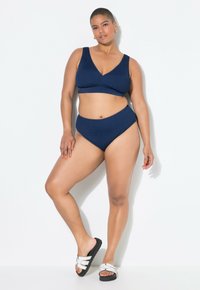 Tmavě modrý bikini set s texturovaným povrchem, obsahující top s V-výstřihem a vysokým pasem. Nosit s černobílými pantofli.