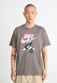 Grå bomullst-skjorte med en pandaillustrasjon som sitter på en sneaker, med en stor rosa Nike-logo i bakgrunnen. Løs passform, korte ermer.