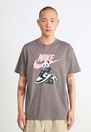 Szürke pamut póló, amelyen egy torna cipőn ülő panda illusztráció látható, a háttérben pedig egy nagy rózsaszín Nike logóval. Engedett szabású, rövid ujjú.
