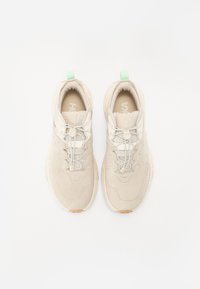 Beige joggesko laget av mesh og semsket skinn, med elastiske lisser, polstret krage og en grønn aksent på hælen.