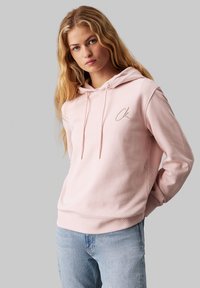 Rosa hettegenser med snøring i hetten og ribbestrikkede mansjetter. Har et lite, brodert "CK"-logo på brystet. Brukes sammen med lyseblå jeans.